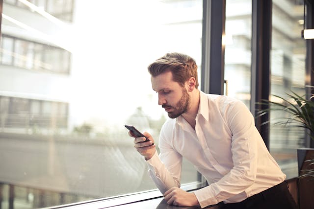 Man in White Dress Shirt Holding Phone Near Window to write in his partner's native language